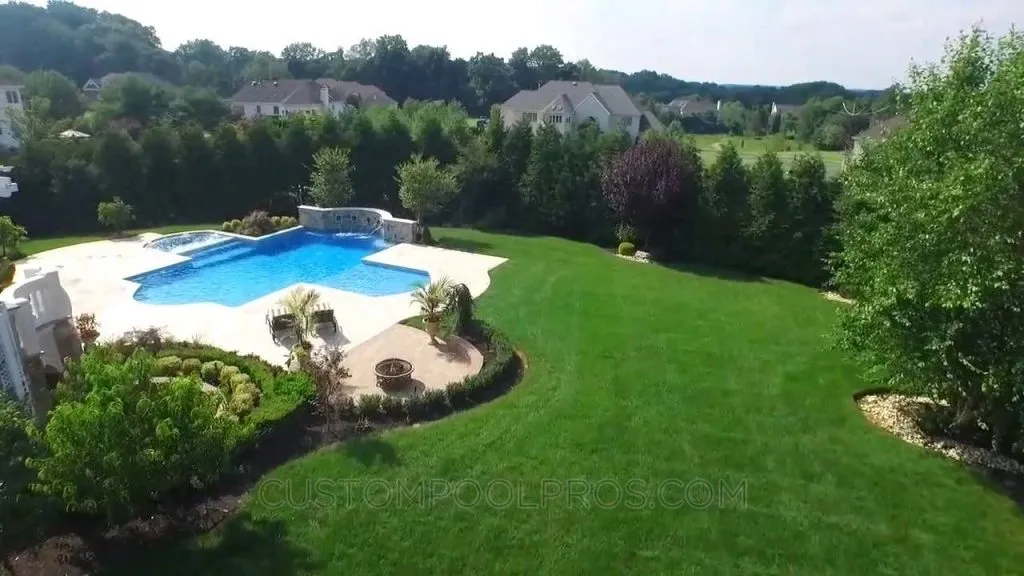 A large backyard with a lush green lawn, landscaped gardens, and a custom-shaped swimming pool perfect for creating pool videos, featuring a stone deck, surrounded by trees and neighboring houses in the background, landscape maintenance