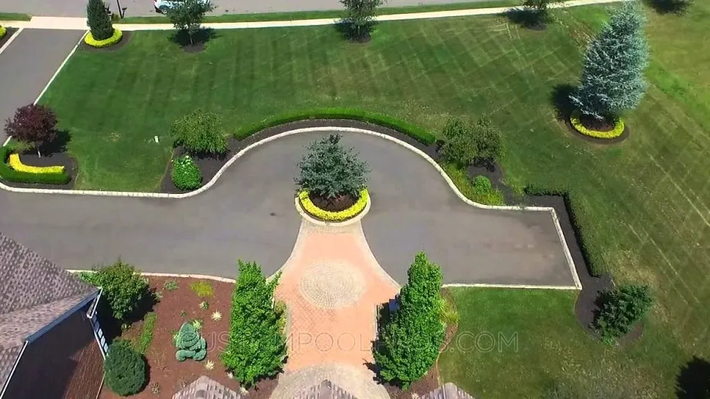 Aerial view of a circular driveway with a central tree, surrounded by neatly trimmed hedges, green lawns, and additional trees in a landscaped yard—perfect for capturing stunning pool videos, landscaping design