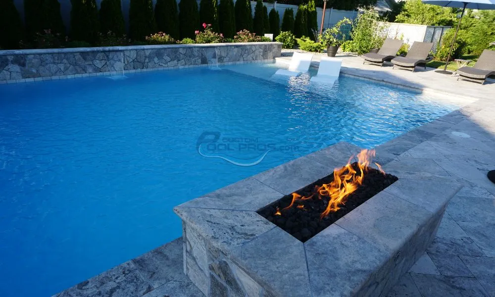 Luxury swimming pools and backyard pool with a stone fire pit and modern lounge chairs. The crystal-clear water reflects the sunlight, surrounded by lush greenery and elegant landscaping, creating a serene outdoor oasis. Ideal for relaxation and entertaining, swimming pool installation new jersey