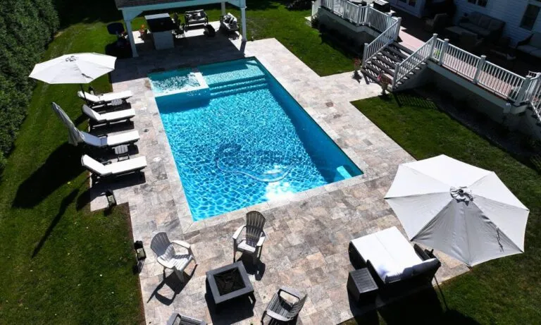 Aerial view of a beautiful backyard pool area featuring a sparkling blue swimming pool surrounded by stone tiles. White lounge chairs and umbrellas provide shade, while a fire pit adds a cozy touch. The lush green lawn enhances the outdoor relaxation space, perfect for summer gatherings, luxury swimming pools