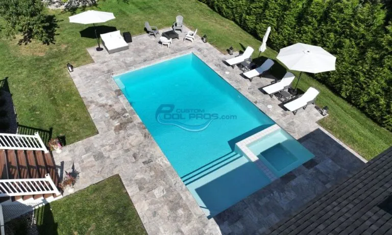 Aerial view of a modern backyard featuring a crystal-clear swimming pool surrounded by elegant stone decking, lounge chairs, and umbrellas, with lush green landscaping in the background, inground pool installation