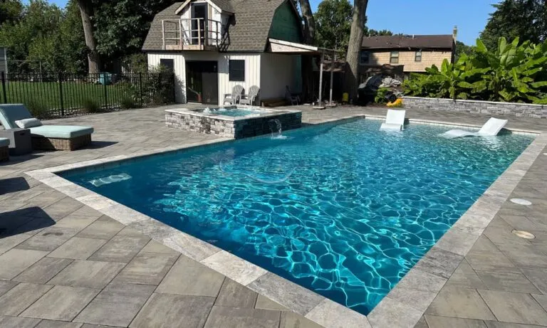 Backyard swimming pool with crystal clear water, surrounded by stone patio and landscaping, featuring a small waterfall and a nearby house with a sloped roof. Ideal for outdoor relaxation and leisure activities, pool builders, swimming pool companies nj