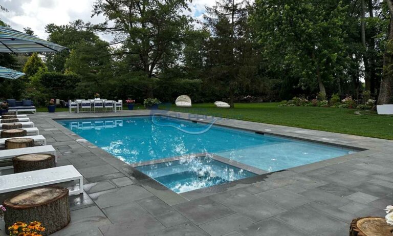Backyard swimming pool surrounded by green grass and trees, featuring a modern design with a stone patio and lounge chairs. Ideal for relaxation and outdoor gatherings, swimming pool installation new jersey