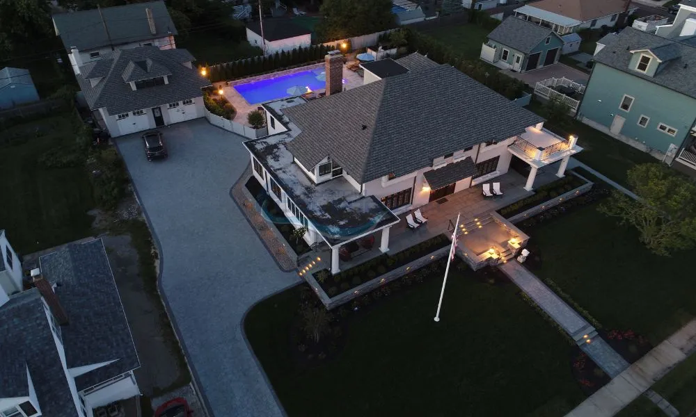Aerial view of a spacious luxury home with a swimming pool, well-lit outdoor areas, and a landscaped yard, surrounded by residential properties at dusk, New Jersey Pool Companies
