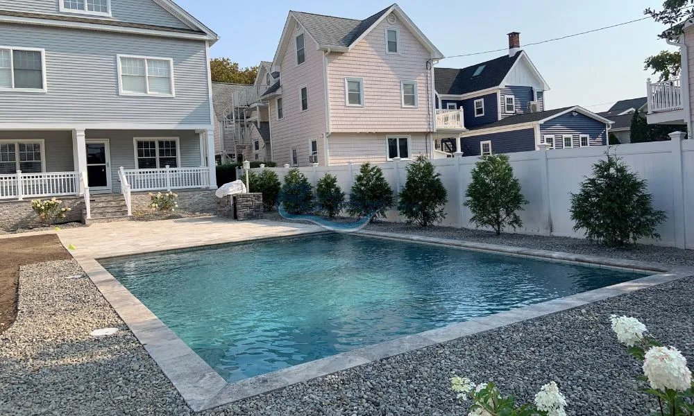 Luxury pools, modern backyard featuring a rectangular swimming pool surrounded by gravel and landscaped greenery, with a two-story house in the background. The scene showcases a relaxing outdoor space ideal for leisure and entertainment, pool contractors