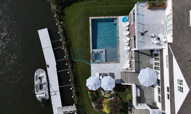 Aerial view of a luxurious backyard featuring a swimming pool, lounge chairs, and umbrellas, adjacent to a docked boat along a serene waterway, inground swimming pool, inground pool builders, pool builders nj