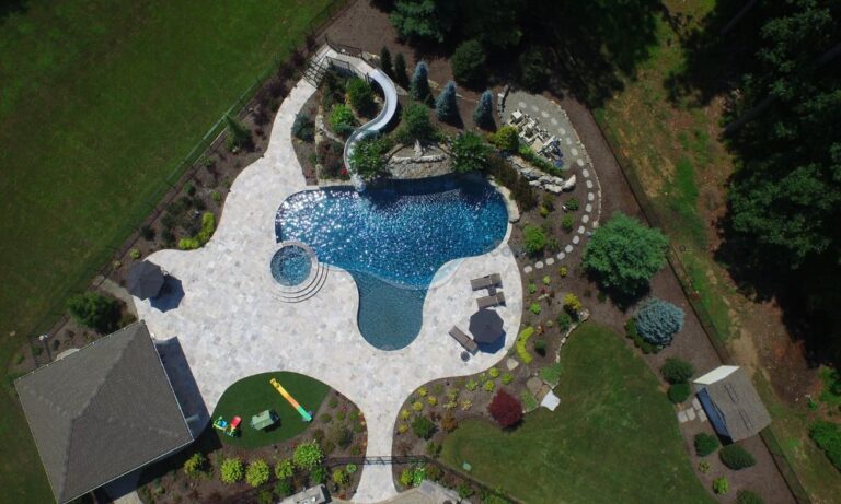 Aerial view of a luxurious backyard with a sparkling blue swimming pool, featuring a water slide and surrounded by landscaped gardens, patio furniture, and a small gazebo. The area is designed for relaxation and recreation, showcasing vibrant greenery and decorative stone pathways, pool builders nj