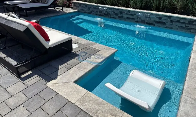 Luxury backyard pool area featuring a crystal-clear swimming pool with water features, surrounded by elegant pavers and modern lounge chairs. A white floating lounge chair is visible in the water, creating a relaxing atmosphere for outdoor enjoyment, swimming pool contractors